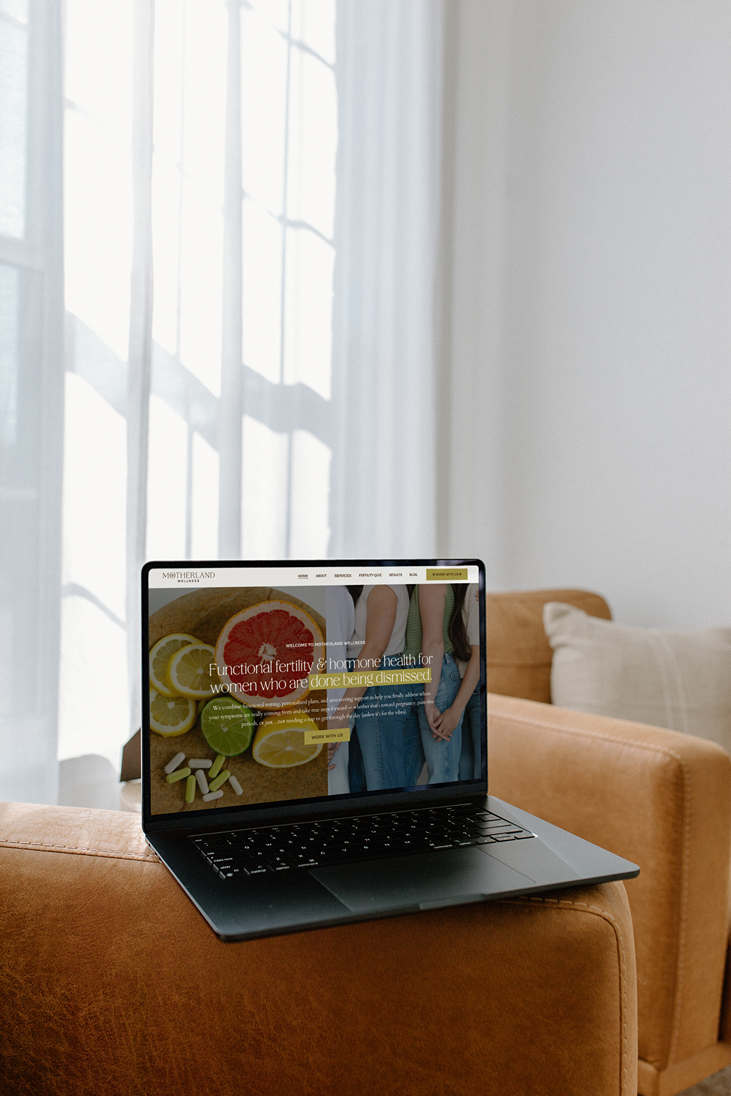 a laptop displaying the Motherland Wellness website sits on a leather armchair in a sun-drenched room